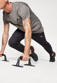 Des barres de pompes noires sur une surface blanche, tenues par une personne portant un t-shirt gris et un pantalon noir, avec des chaussures noires aux accents rouges.
