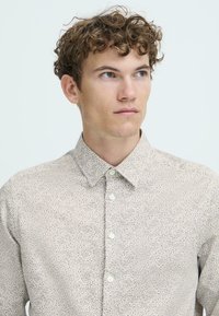 Beige skjorte med knapper, preget av et lite, gjentatt blomsterdesign, klassisk krage og standard knappelukking, med en glatt stofftekstur.