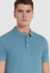 Polo en bleu clair, fabriqué en tissu texturé. Il comporte un col, une patte de boutonnage à trois boutons et un petit logo orange sur la poitrine.