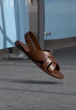 Brown leather sandal with woven straps and ankle buckle, suspended mid-air above paved ground near a blue barrier.