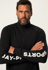Schwarzes Sportoberteil mit langen Ärmeln und Reißverschlusskragen. Mit weißem Text auf den Ärmeln, der "JAY-P SPORTS" liest. Weicher Stoff mit taillierter Passform.