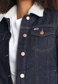 Denim jacket in dark blue with a white fleece collar. Features copper buttons, front pockets, and contrasting stitching details.