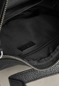 Interiér černé kožené kabelky s hladkou texturou, obsahující kapsu na zip a malou černou štítek, vše na pozadí tmavého látkového podšívky.