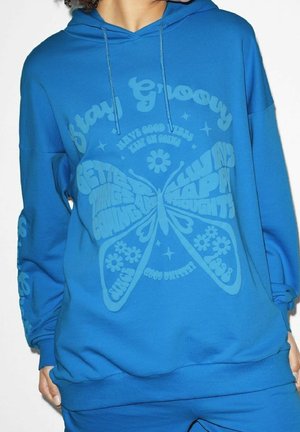 Osoba nosząca niebieską bluzę z kapturem z grafiką motyla i pozytywnymi hasłami takimi jak „Stay Groovy”, „Better Things Are Coming” oraz „Always Happy Thoughts.”