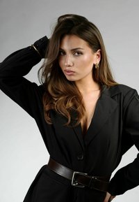 Blazer negro con escote pronunciado, ceñido por un cinturón de cuero marrón. La modelo lleva pendientes de aro dorados y tiene el pelo largo y ondulado.