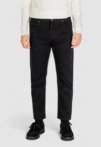 Pantaloni in velluto a coste neri con una vestibilità dritta, tasche anteriori e una texture sottile, indossati con scarpe nere e un maglione di colore chiaro.