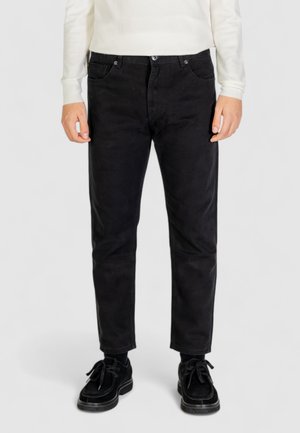 Pantaloni in velluto a coste neri con una vestibilità dritta, tasche anteriori e una texture sottile, indossati con scarpe nere e un maglione di colore chiaro.