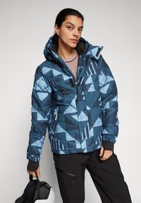 Giubbotto trapuntato blu e nero con un motivo geometrico, dotato di chiusura lampo anteriore, cappuccio e polsini elasticizzati. Abbinato a pantaloni neri.