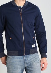 Marineblauwe bomberjack met een geribbelde grijze kraag, koperen rits en twee zijzakken. Voorzien van een aansluitende tailleband en een stoffen label aan de voorkant.