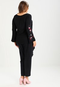 Haut noir en maillot avec broderie florale sur les manches, accompagné d'un pantalon noir ample et de chaussures à talons hauts rose clair.