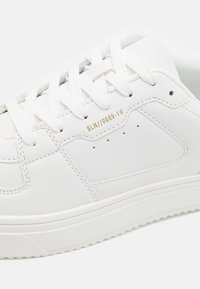 Hvide læder sneakers med flade snørebånd, guldpræget label "BLN//0089-10," rund tå og struktureret gummisål med zigzag mønster.