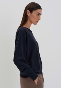 Marineblauer Pullover mit rundem Halsausschnitt und lockerer Passform. Lange Ärmel, minimale Nähte und eine weiche Textur. Kombiniert mit hellen, neutralen Hosen.
