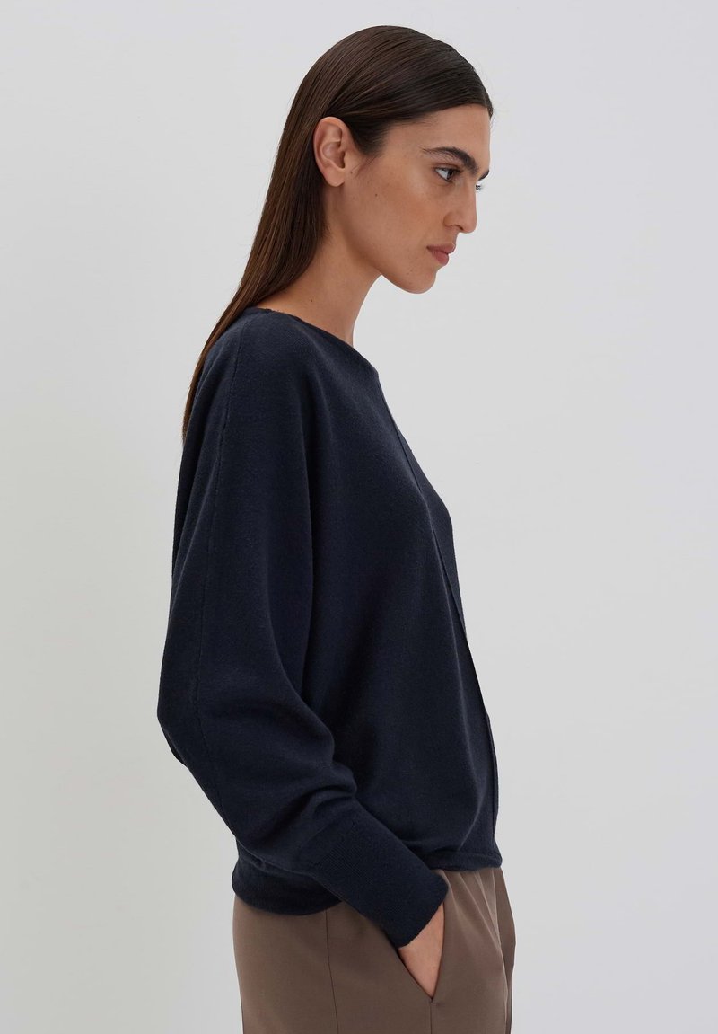 Marineblauer Pullover mit rundem Halsausschnitt und lockerer Passform. Lange Ärmel, minimale Nähte und eine weiche Textur. Kombiniert mit hellen, neutralen Hosen.