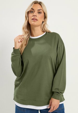 Femme blonde aux cheveux lâches et ondulés portant un sweat-shirt à manches longues vert olive par-dessus une chemise blanche et un jean bleu sur un fond uni.