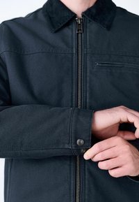 Chaqueta oscura y texturizada con cierre frontal de cremallera, puños abotonados y cuello. Presenta un pequeño bolsillo en el pecho y un acabado suave.
