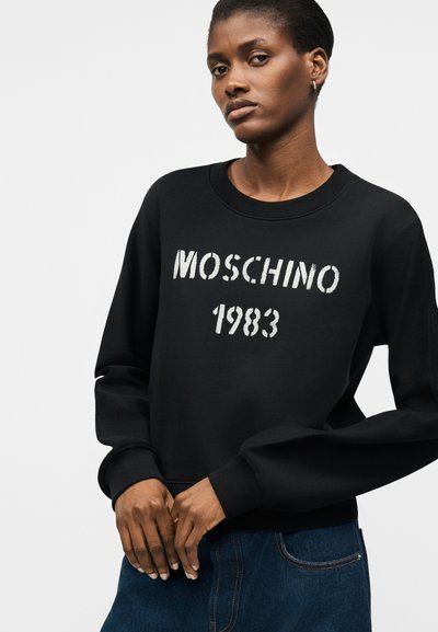 Γυναίκα που φοράει μαύρο φούτερ Moschino 1983 και μπλε τζιν, με τα χέρια σταυρωμένα μπροστά, κοιτώντας ελαφρώς στο πλάι.