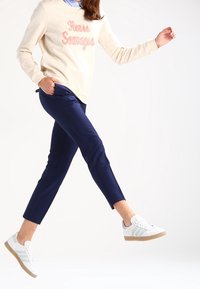 Beige tröja med "Fais Savages" i texturerad rosa text, marinblå tajta byxor och vita sneakers med ljusgröna accenter och gummisulor.
