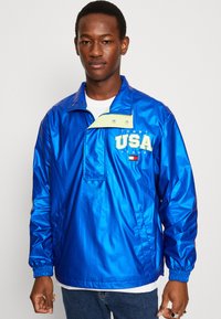 Blå nylon genserjakke med høy krage, trykknapplukking, frontlomme og "USA" grafikk i neongul. Elastiske mansjetter, lett tekstur.