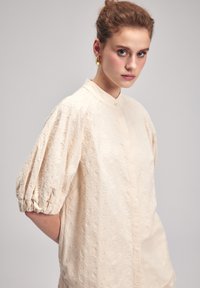 Cremfarbige Bluse mit Puffärmeln, besticktem Blumenmuster, hohem Kragen und Knopfverschluss. Strukturierter Stoff mit zarten Details.