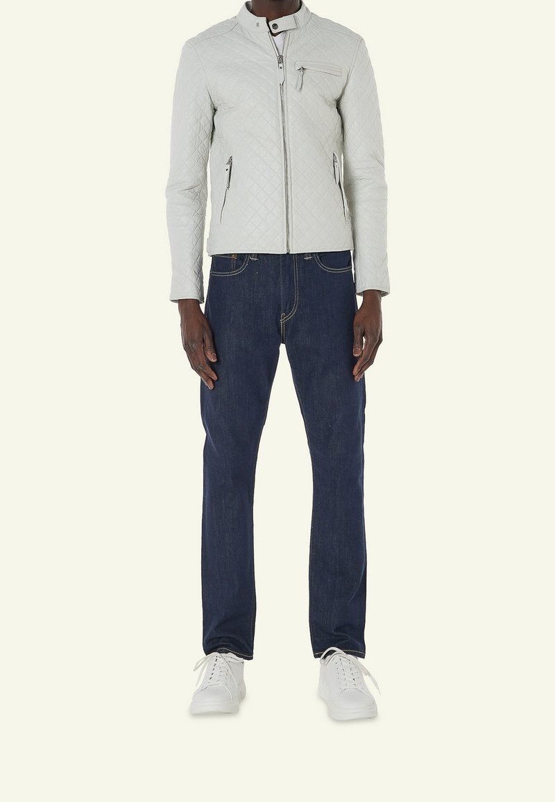 Personne portant une veste matelassée gris clair avec col montant, jean bleu foncé et baskets blanches, debout devant un fond uni.