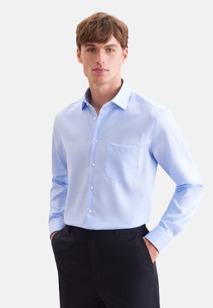 Jeune homme portant une chemise bleu clair à boutons avec une poche poitrine et un pantalon noir, debout les mains dans les poches devant un fond uni.