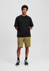 Tricou negru texturat cu modele geometrice, purtat cu pantaloni scurți khaki. Modelul poartă adidași negri și albi cu accente verzi.
