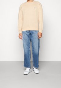 Felpa beige testurizzata con polsini e orlo a coste, abbinata a jeans dritti blu sbiaditi e sneakers bianche con accenti grigi.