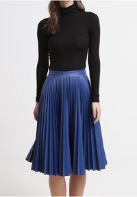 Plisserad blå kjol i slät tyg, med en åtsittande midja och en A-linjeform. Svart polotröja bärs ovanför kjolen.