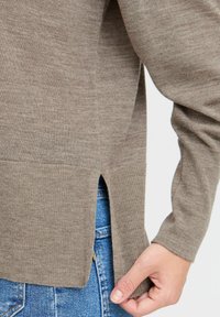 Pull marron clair avec une texture douce, doté d'une fente sur le côté et de poignets côtelés, assorti à un jean en denim bleu.