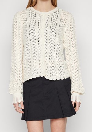 Femme portant un cardigan blanc en maille à manches longues boutonné avec des bords festonnés et une mini-jupe noire plissée.