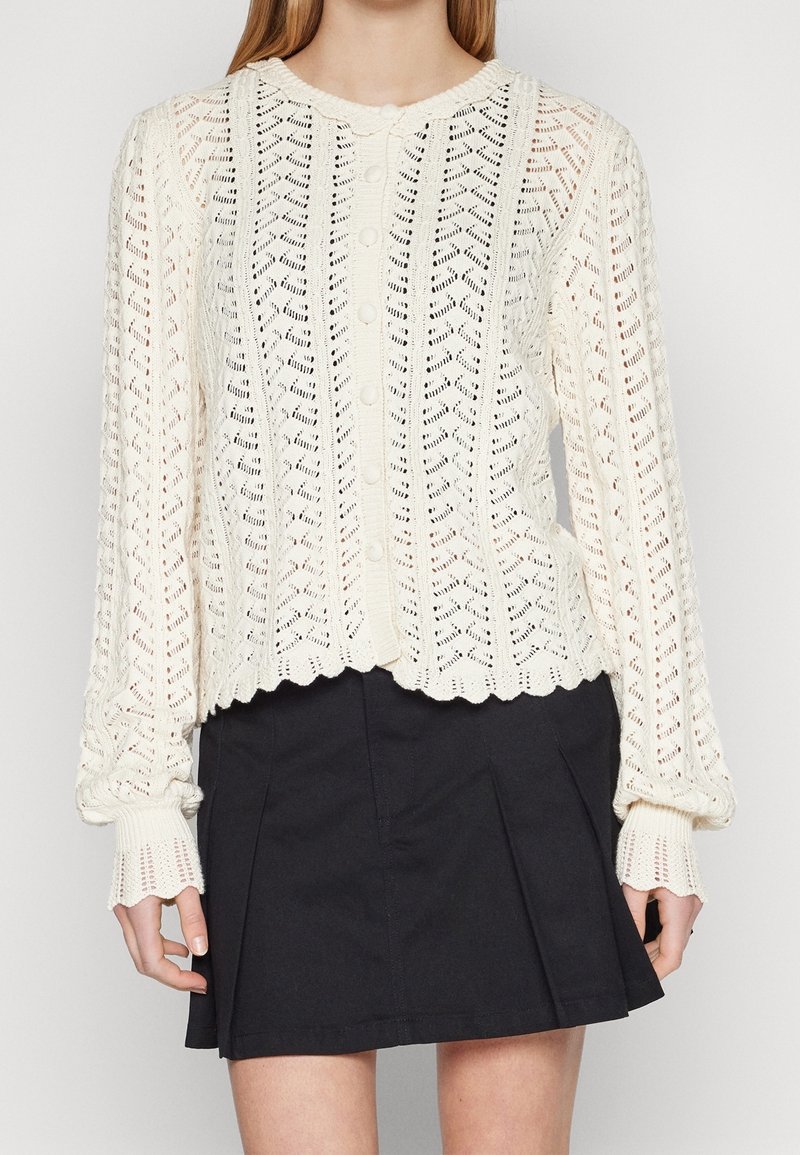 Femme portant un cardigan blanc en maille à manches longues boutonné avec des bords festonnés et une mini-jupe noire plissée.