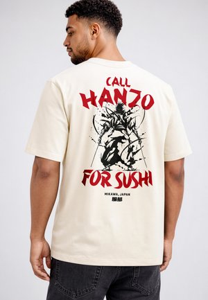 Mann trägt cremefarbenes T-Shirt mit rotem Schriftzug „Call Hanzo For Sushi“ und schwarzer Tuschezeichnung eines Samurais auf dem Rücken.