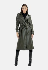 Olivgrön trenchcoat i konstläder, dubbelknäppt med skärp i midjan, med vida slag och midjelångt snitt. Svarta knähöga stövlar.