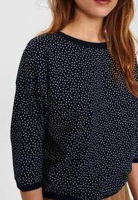 Marineblauer Pullover mit weißen Tupfen, Rundhalsausschnitt und dreiviertellangen Ärmeln. Weicher Stoff und geripptes Saumdetail.