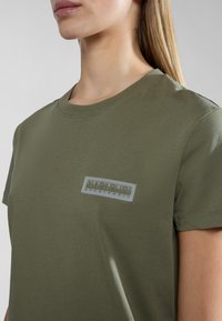Oliivinvihreä puuvillainen t-paita, jossa on pyöreä pääntie ja vasemmassa rinnassa suorakulmion muotoinen harmaa logokangas. Sujuva pinta, lyhyet hihat.