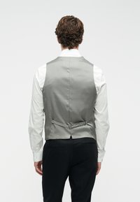 Mees, kelles on valge särg, hõbedane reguleeritava seljaosaga vöö, ja mustad püksid, seisab tagasi pööratud suundudes lihtsa valge tausta poole.