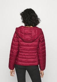 Rød dunjakke med hætte, med vandret quiltning og glat tekstur. Tætsiddende ærmer og kant. Sorte bukser er iført.