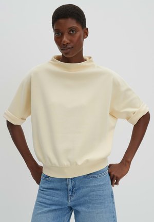 Vrouw met kort haar die een crèmekleurige top met korte mouwen en opgerolde boorden draagt, en een blauwe spijkerbroek, staand met handen in de zij.