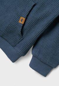 Maglione blu lavorato a maglia con texture a coste, maniche con risvolto e un etichetta in tessuto marrone con un dettaglio del logo.