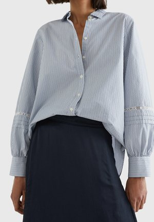 Vrouw draagt een loszittend, blauw-wit gestreept overhemd met opgeblazen mouwen en een donker marineblauwe rok tegen een effen achtergrond.