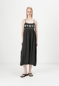 Abito maxi nero con spalline sottili, ricamo floreale bianco e una vestibilità ampia. Indossato con sandali neri, caratterizzati da una trama fluida.