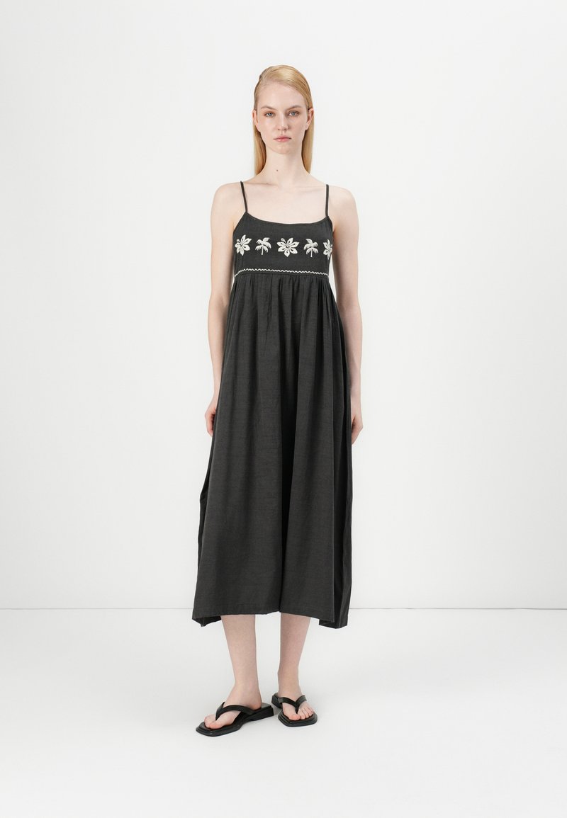 Abito maxi nero con spalline sottili, ricamo floreale bianco e una vestibilità ampia. Indossato con sandali neri, caratterizzati da una trama fluida.