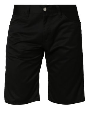 Grauer Pullover mit Raglanärmeln, strukturierter Stoff und gerippte Bündchen, kombiniert mit schwarzen Shorts und schwarzen Sneakern. Sonnenbrille in der Hand.