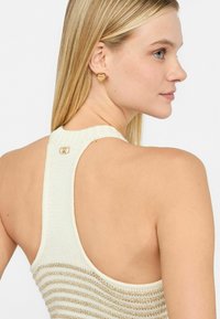 Top in maglia con schiena scoperta, caratterizzato da strisce verticali dorate e un dettaglio con bottone sul collo. Orecchini a forma di cuore in oro sull'orecchio della modella.