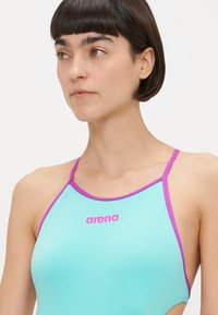 Τουρκουάζ μαγιό με μοβ λουριά, που διαθέτει λογότυπο σε ροζ. Λείο υλικό και αθλητικός σχεδιασμός κατάλληλος για κολύμβηση.