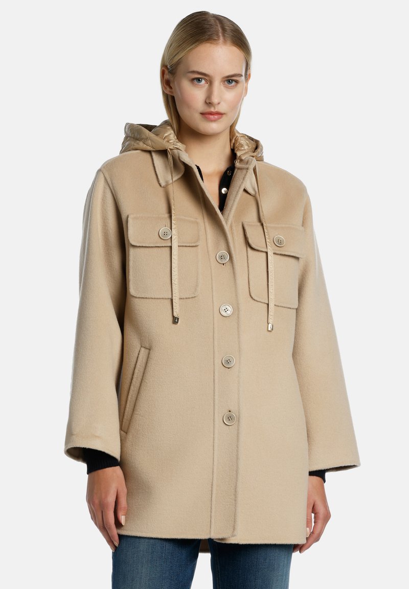 Cappotto beige con cappuccio realizzato in tessuto morbido, con due tasche sul petto con bottone, cappuccio regolabile e bottini frontali. Presenta una vestibilità comoda e tasche laterali.