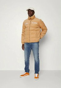 Homem a usar uma jaqueta puffer bege com o texto "KARL KANI", jeans azuis, ténis laranjas e boné branco, posando contra um fundo simples.