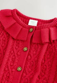 Rød strikket cardigan med et struktureret kabeldesign, der har en rund krave, tre træknapper og et mærke med teksten "NEXT baby."