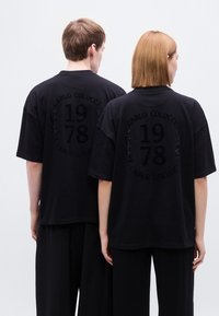 Μαύρα υπερμεγέθη μπλουζάκια με ανάγλυφο κείμενο και σχέδια δάφνης στην πλάτη, που απεικονίζουν το "CARLO COLUCCI 1978." Λείο βαμβακερό ύφασμα.