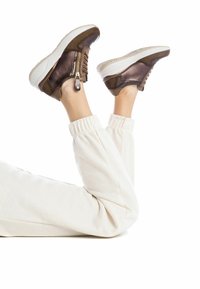 Zapatillas de cuero marrón con un zipper lateral. Presentan una suela blanca con acentos texturizados. Se llevan con pantalones jogger claros y fruncidos.